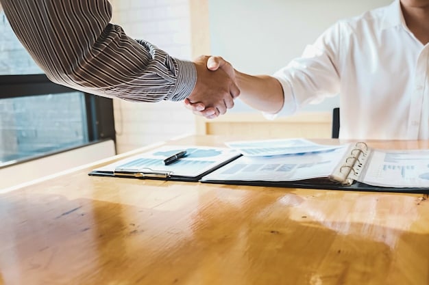 Two hands shaking over a table with documents and a pen, representing agreement and official processes for financial assistance. The setting is bright and professional.