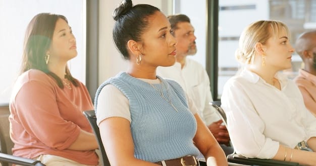 A diverse group of adults participating in a job training workshop, with a facilitator at the front. They are engaged in a discussion, some taking notes on laptops or notebooks, signifying active learning and career development.